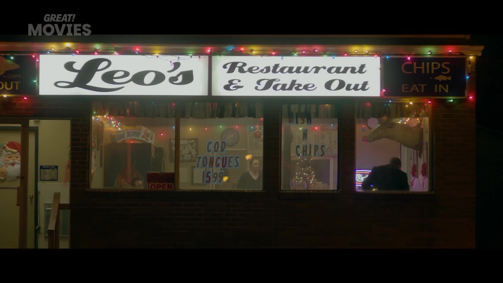 A man in a suit stands behind the counter of Leo's Restaurant & Take Out, illuminated by a neon sign. Christmas lights twinkle above the storefront, casting a festive glow on the chilly evening.