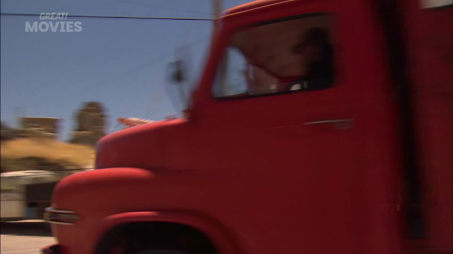 A bright red lorry is rumbling past. The driver's silhouette is visible through the window, and the vehicle appears to be on a UK road.