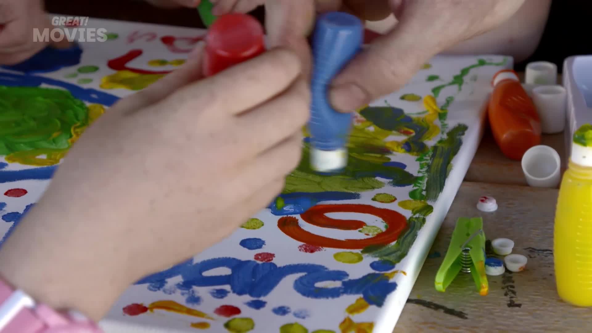 A child's hand squeezes blue paint from a bottle onto a canvas. Another hand, likely an adult's, guides a red paint bottle, adding to the colourful artwork.
