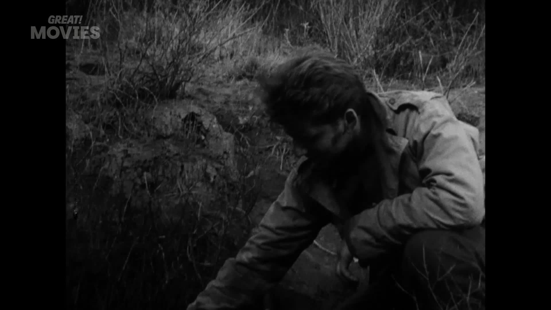A man in a jacket crouches, reaching into the rough, uneven ground. He appears to be examining something in the dirt, his face obscured by shadow.