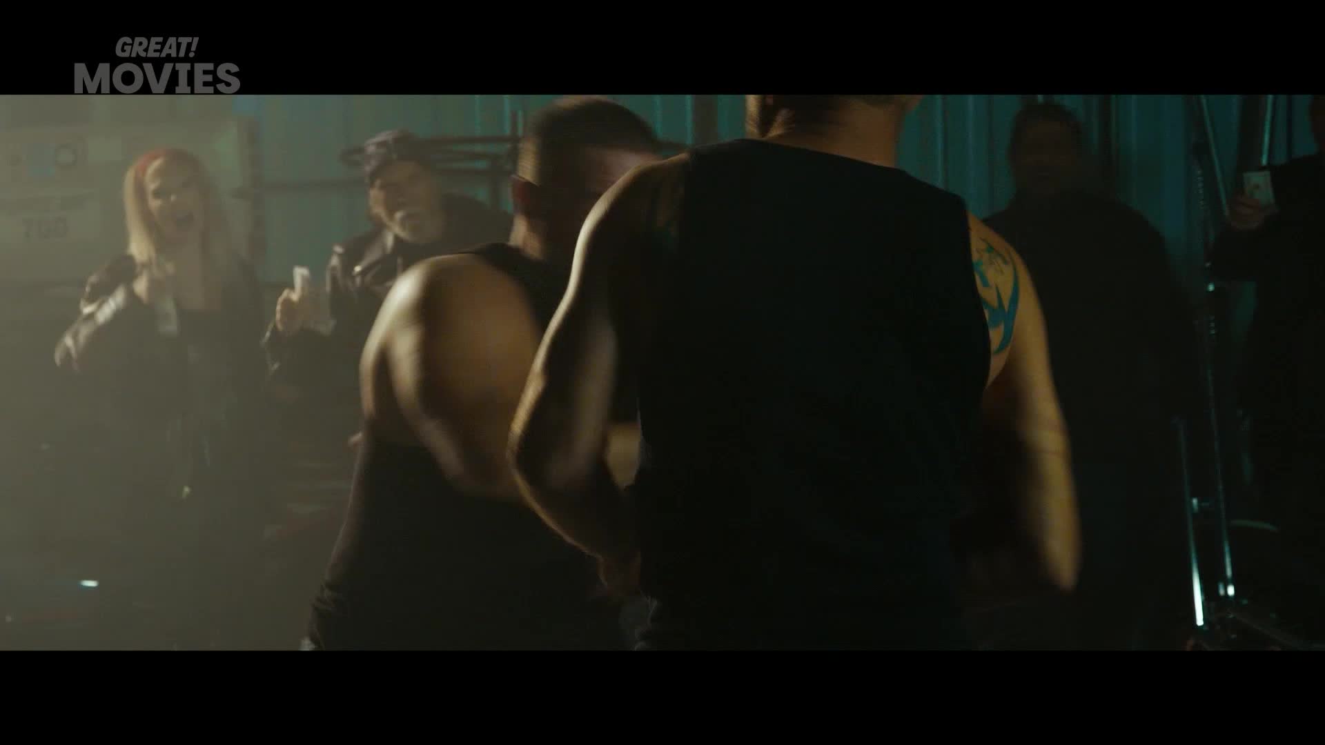 Two men in black tank tops are facing each other, one with a blue tattoo on his arm. A woman with blonde hair and a leather jacket is behind them, laughing.