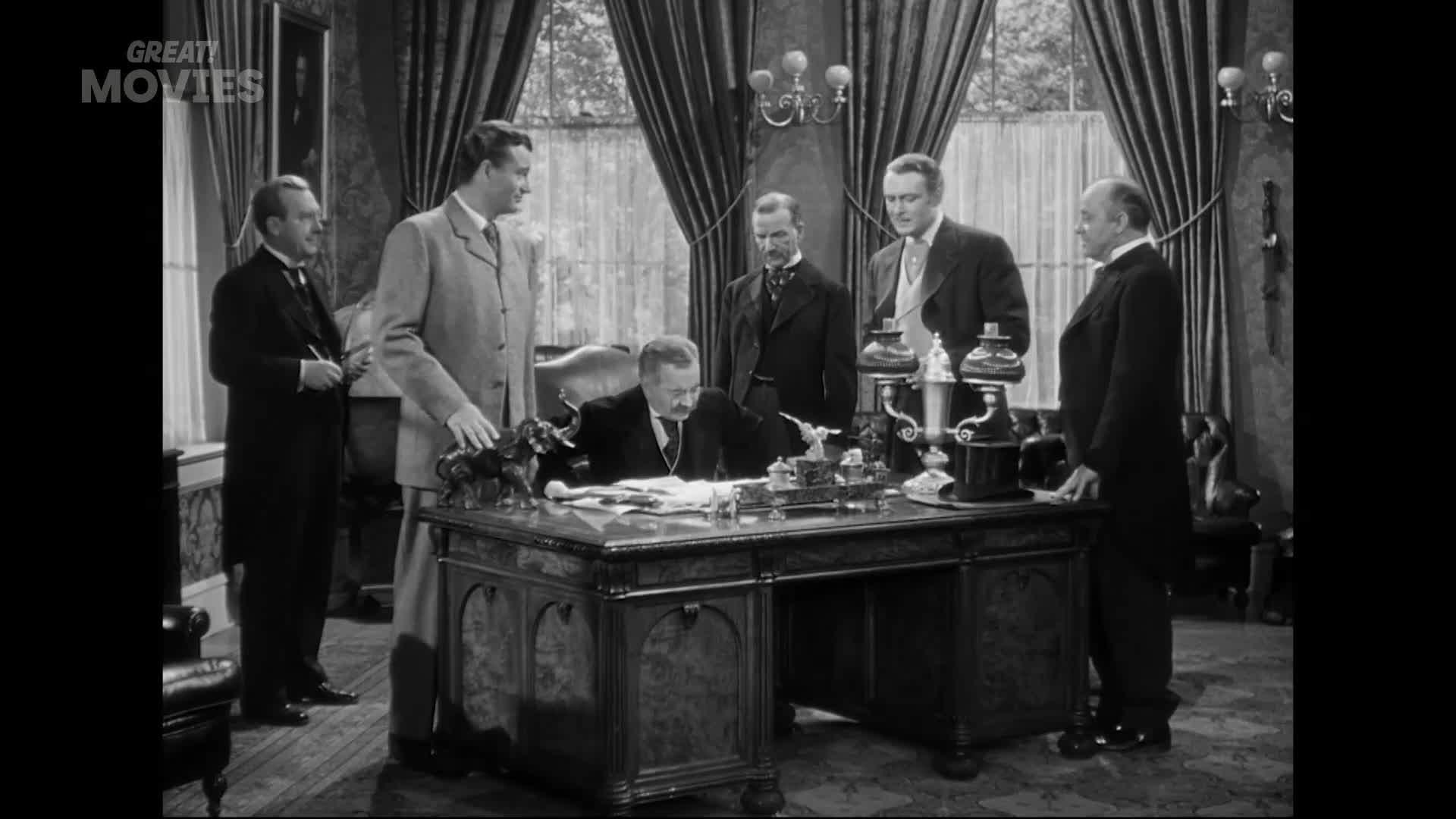 A man in a light suit leans over a large desk, signing papers. Four other men in formal attire stand around him, observing the scene.