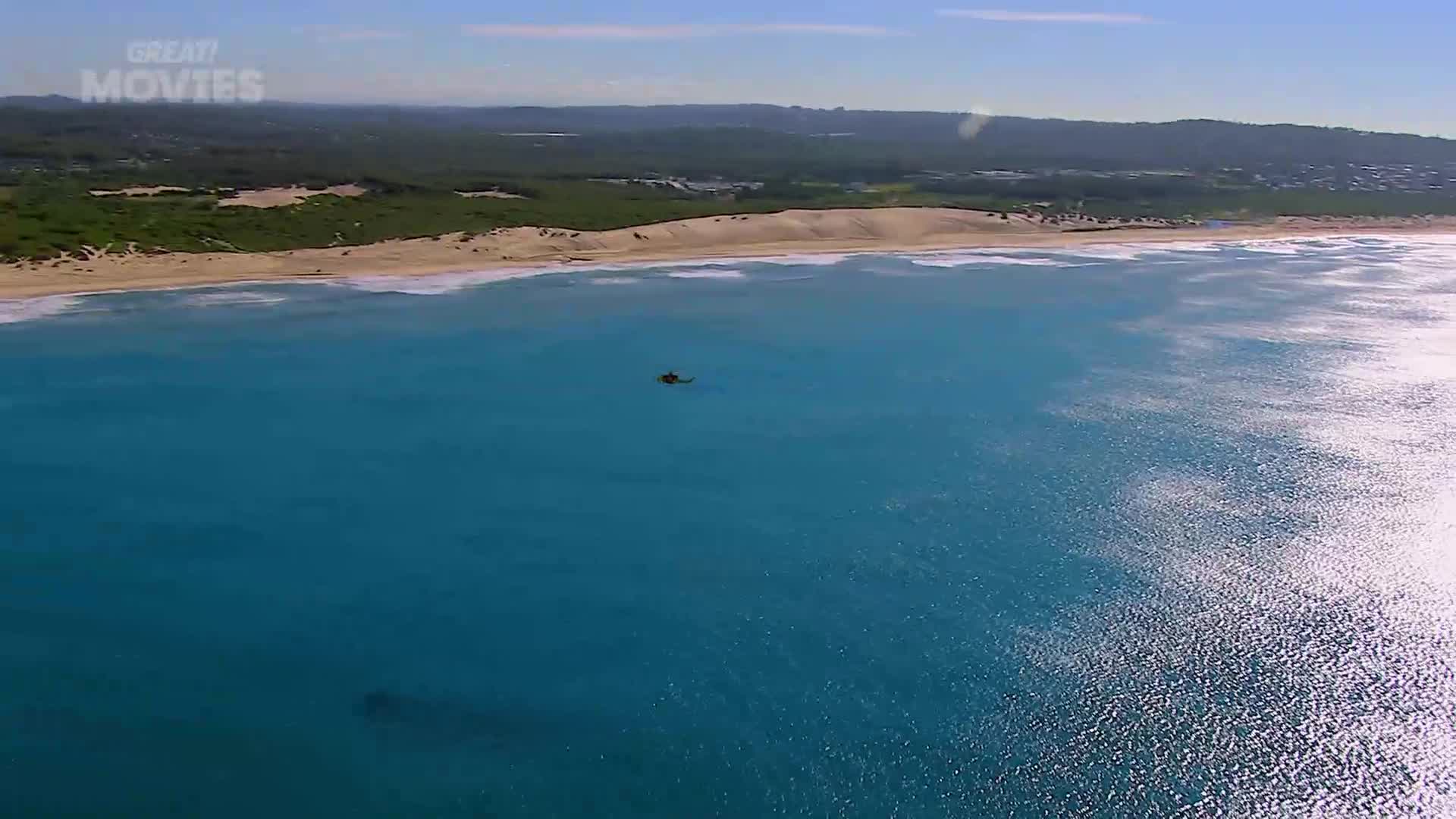 A helicopter hovers over the bright blue sea, not far from a sandy coastline. The waves roll in, creating white lines against the shore.
