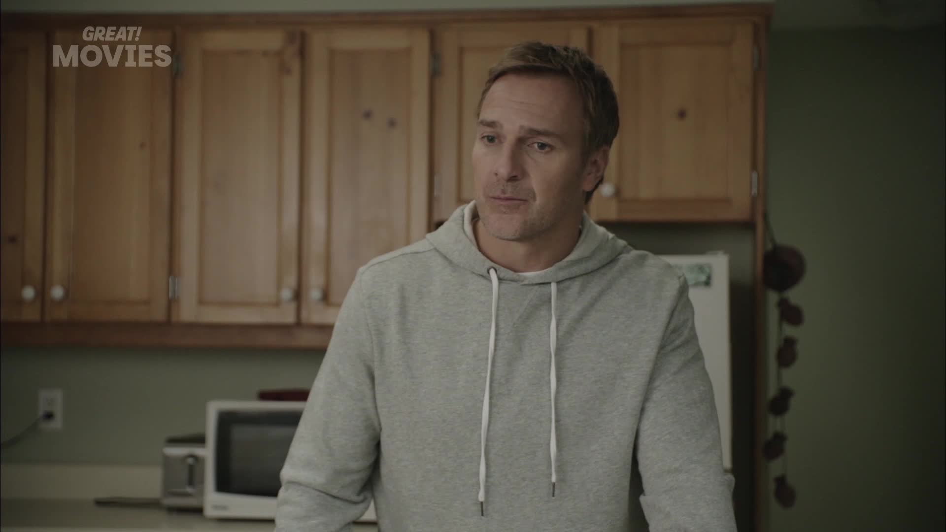 A man in a grey hoodie stands in a kitchen. He looks off to the side, his brow furrowed. The "GREAT! MOVIES" logo is visible in the top left corner.