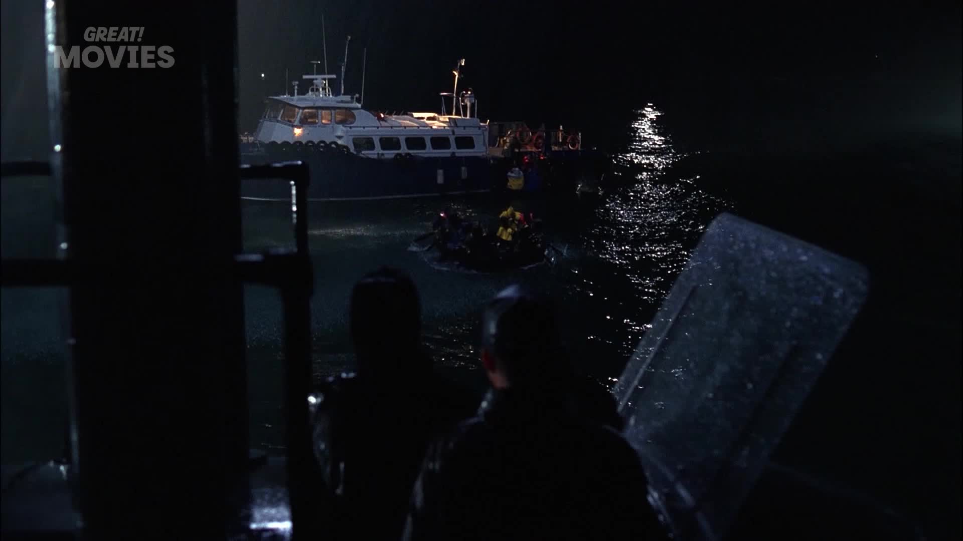 A large boat is docked, its lights reflecting on the dark water. A smaller, overloaded dinghy is being pulled alongside, its occupants huddled together.