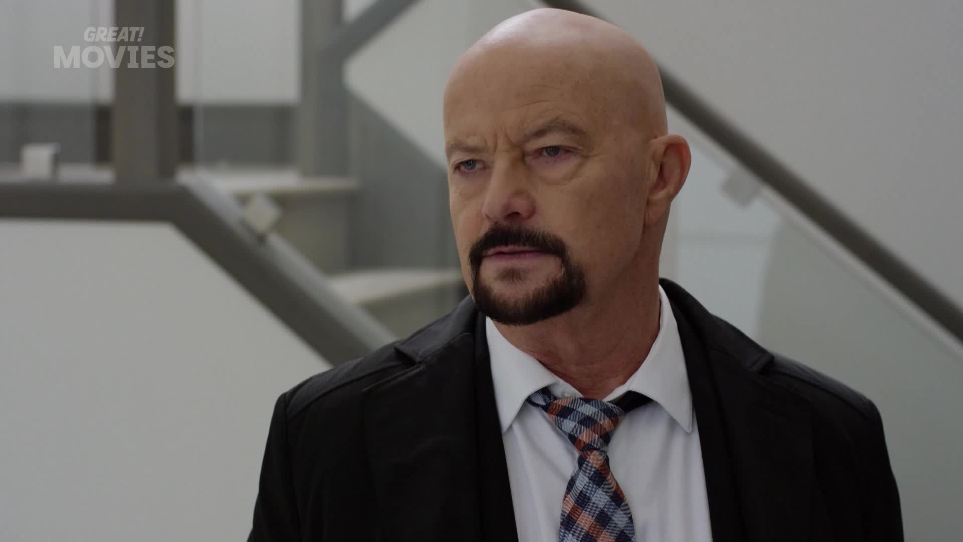 A man with a shaved head and a goatee stands looking off to the side. He wears a dark jacket over a white shirt and a plaid tie.