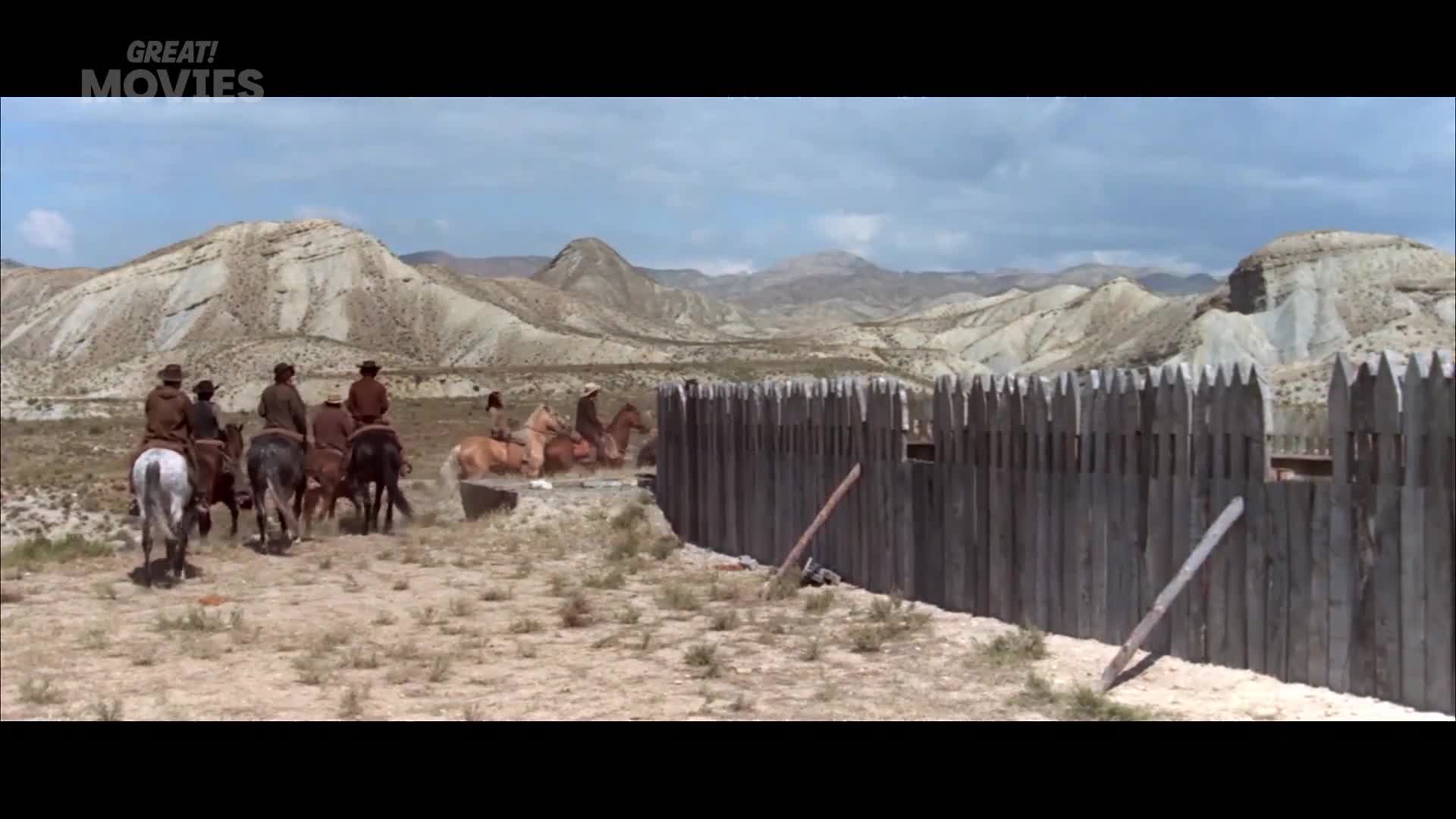 A group of riders approaches a wooden palisade. Their horses kick up dust as they move across the arid landscape.