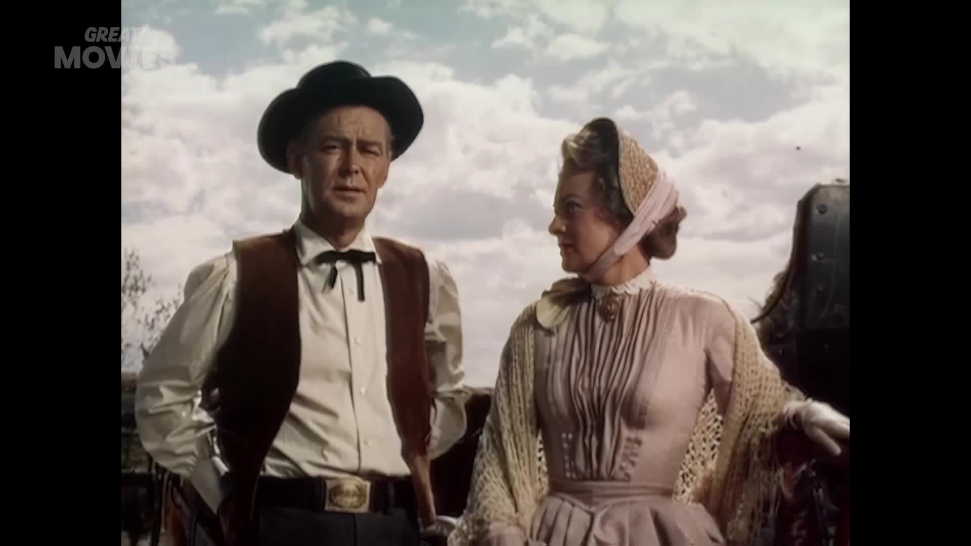 A man in a cowboy hat and vest stands next to a woman in a bonnet and lace shawl. They're both looking off to the side, as if watching something approach.