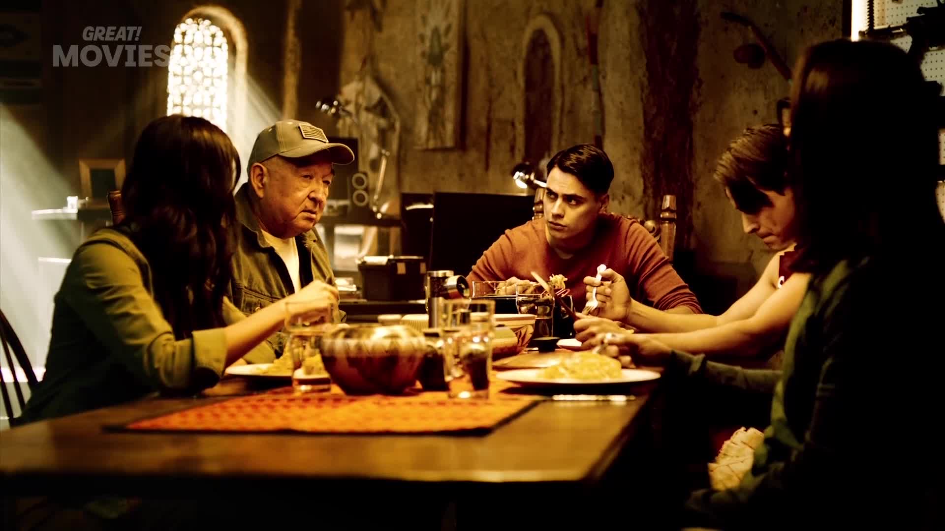A family gathers around a table, their faces illuminated by a single light source. The man in the cap gestures with his fork as he speaks, while the younger man opposite him listens intently.