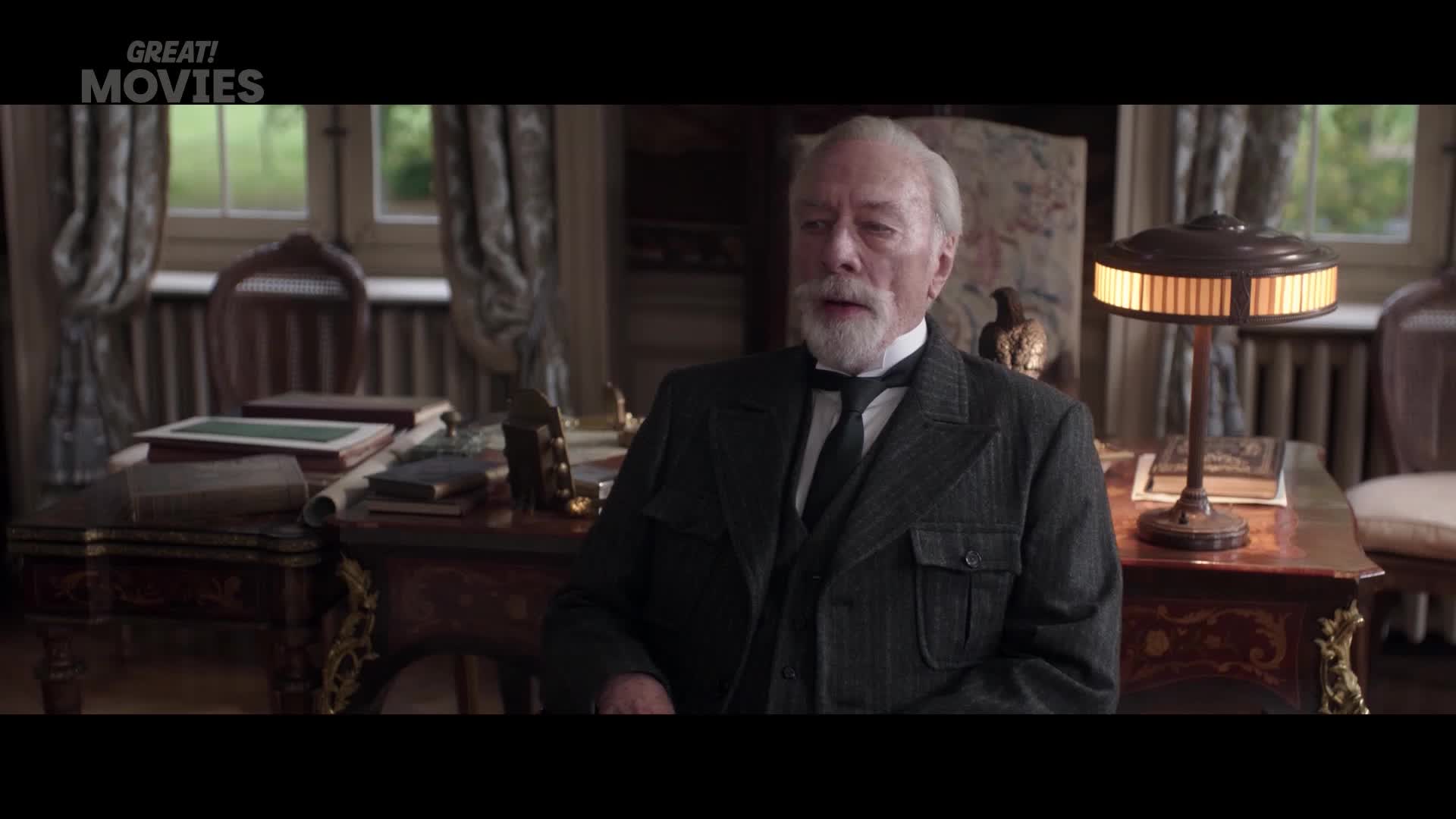 An older gentleman in a dark suit and tie sits at a wooden desk, his gaze fixed forward. A lamp with a ribbed shade casts a warm glow on the surface of the desk, illuminating stacks of books and papers.