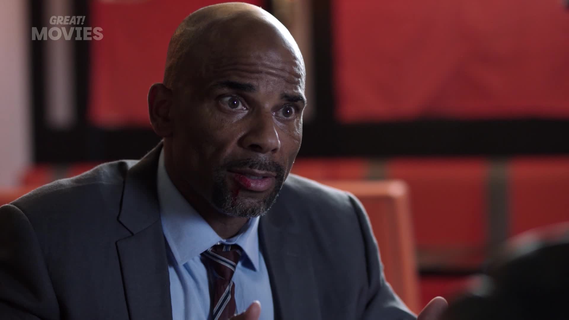 A man in a grey suit and striped tie leans forward, his expression intense. He's speaking with urgency, a small cut visible on his lip.