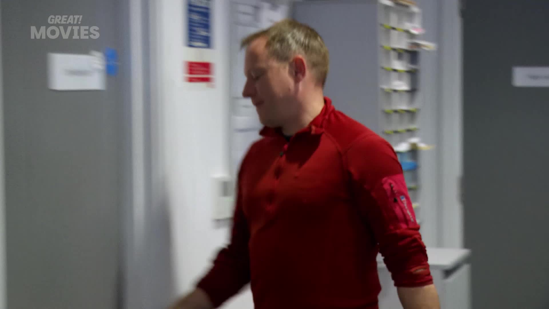 A man in a red top is walking past a door with a "GREAT! MOVIES" logo. He's moving quickly, his arm swinging slightly as he passes.