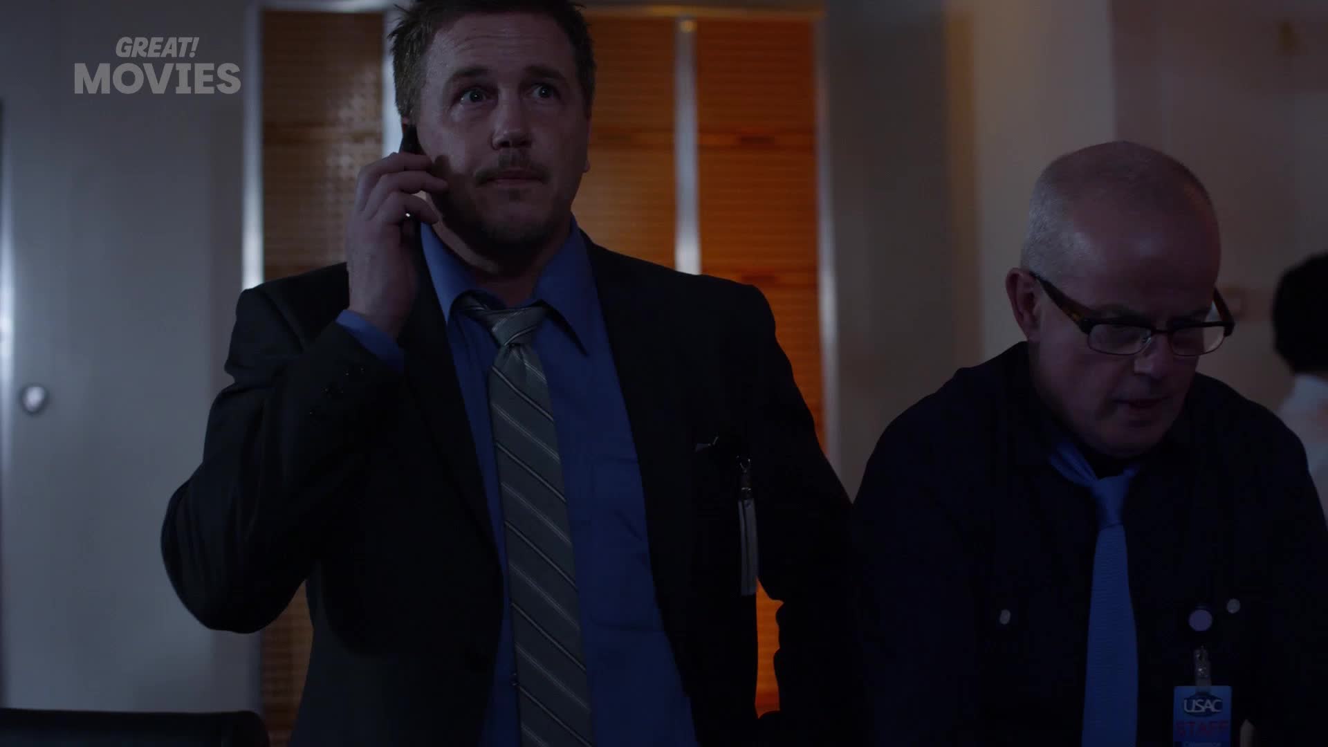 A man in a suit speaks intently on his mobile phone, his gaze fixed upwards. Beside him, another man in a dark shirt and tie looks down, his glasses reflecting the dim light.