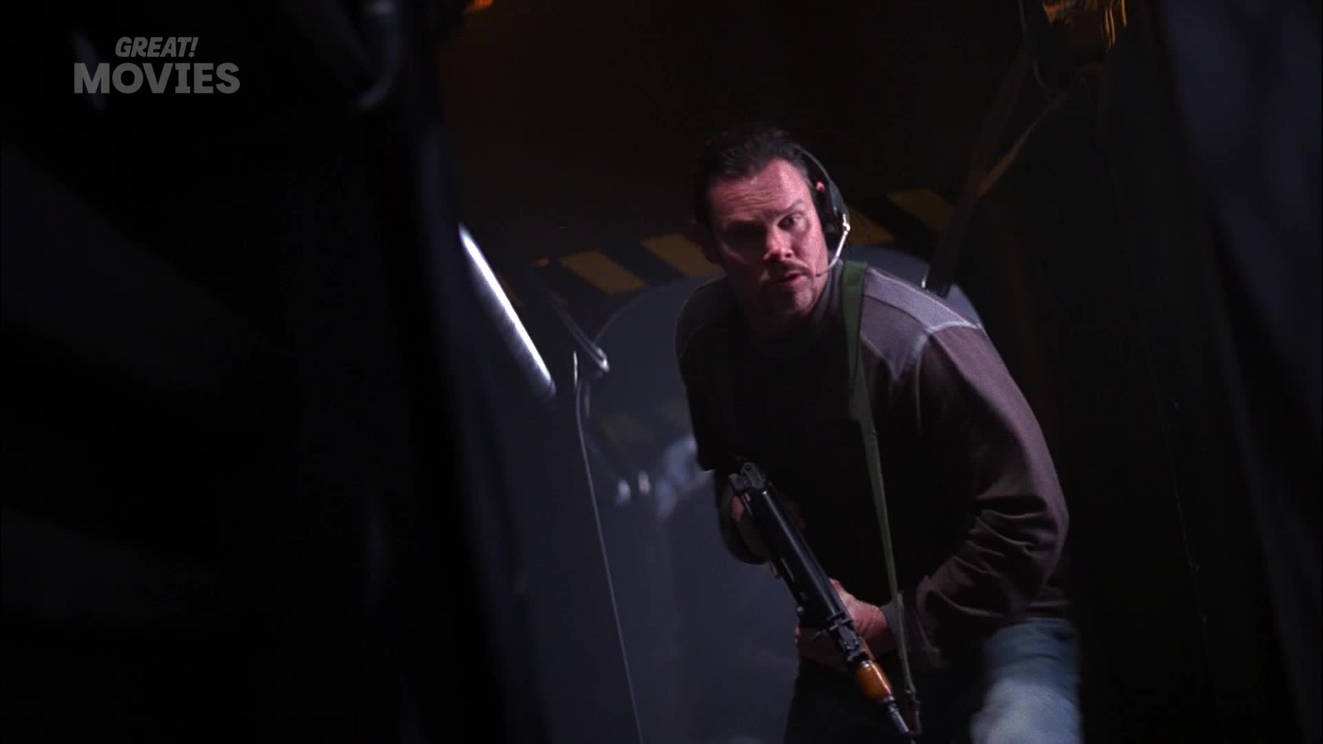 A man with a headset and a rifle crouches, his eyes wide with alarm. He's in a dimly lit, industrial-looking space, possibly a warehouse or a film set.