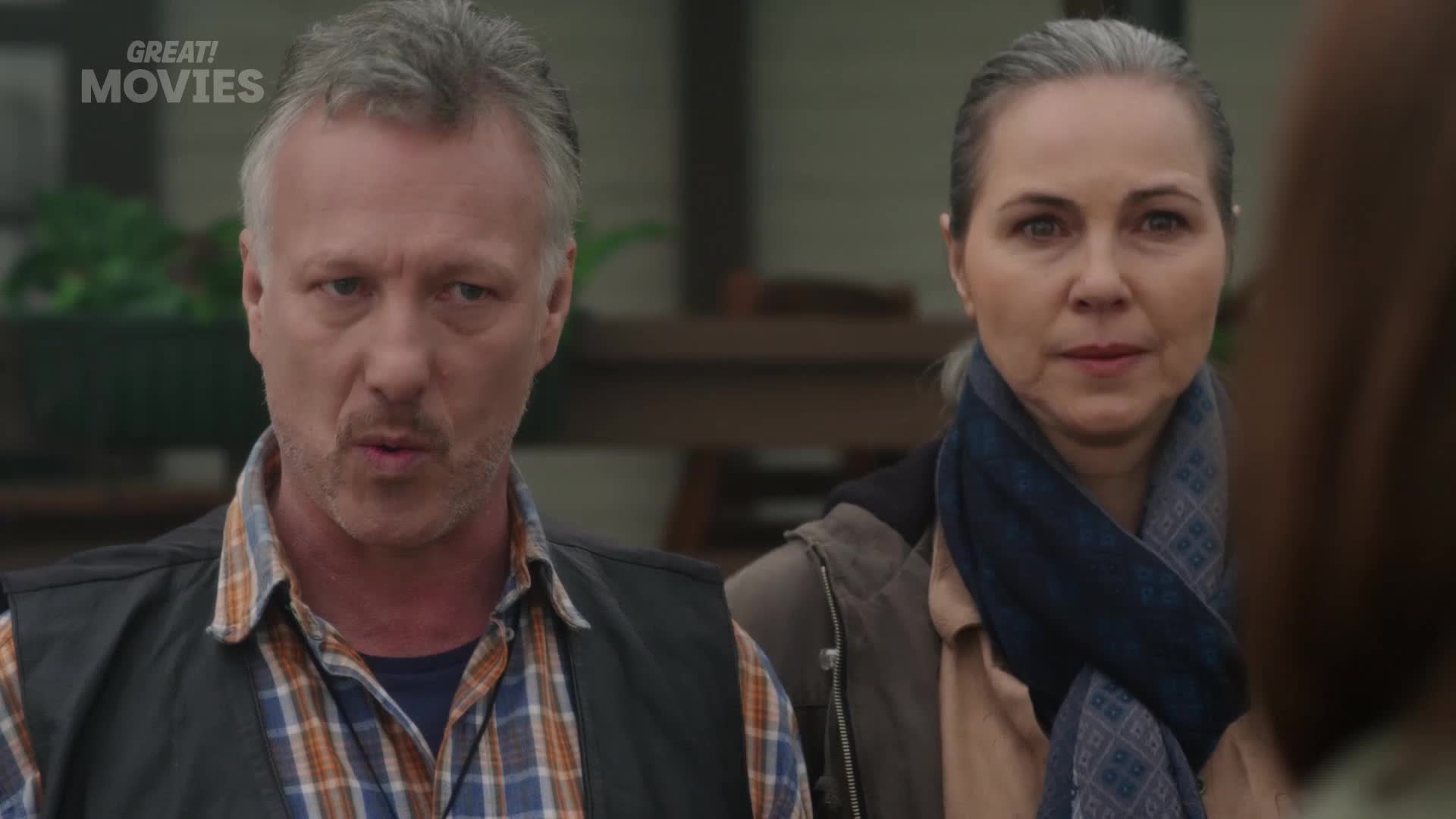 A man with grey hair and a plaid shirt speaks intently, his expression serious. Beside him, a woman with her hair pulled back gazes forward, a patterned scarf around her neck.