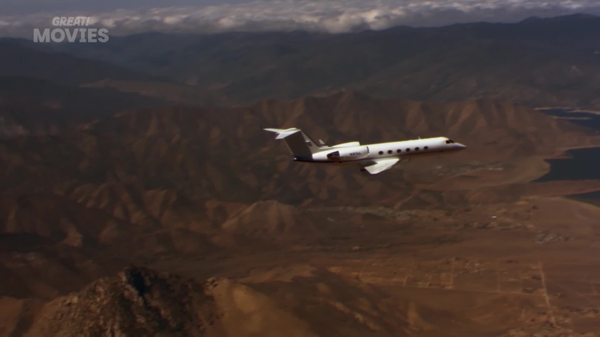 A sleek white jet cruises over rugged, arid terrain. The plane glints in the sun as it flies towards a dark, still body of water.