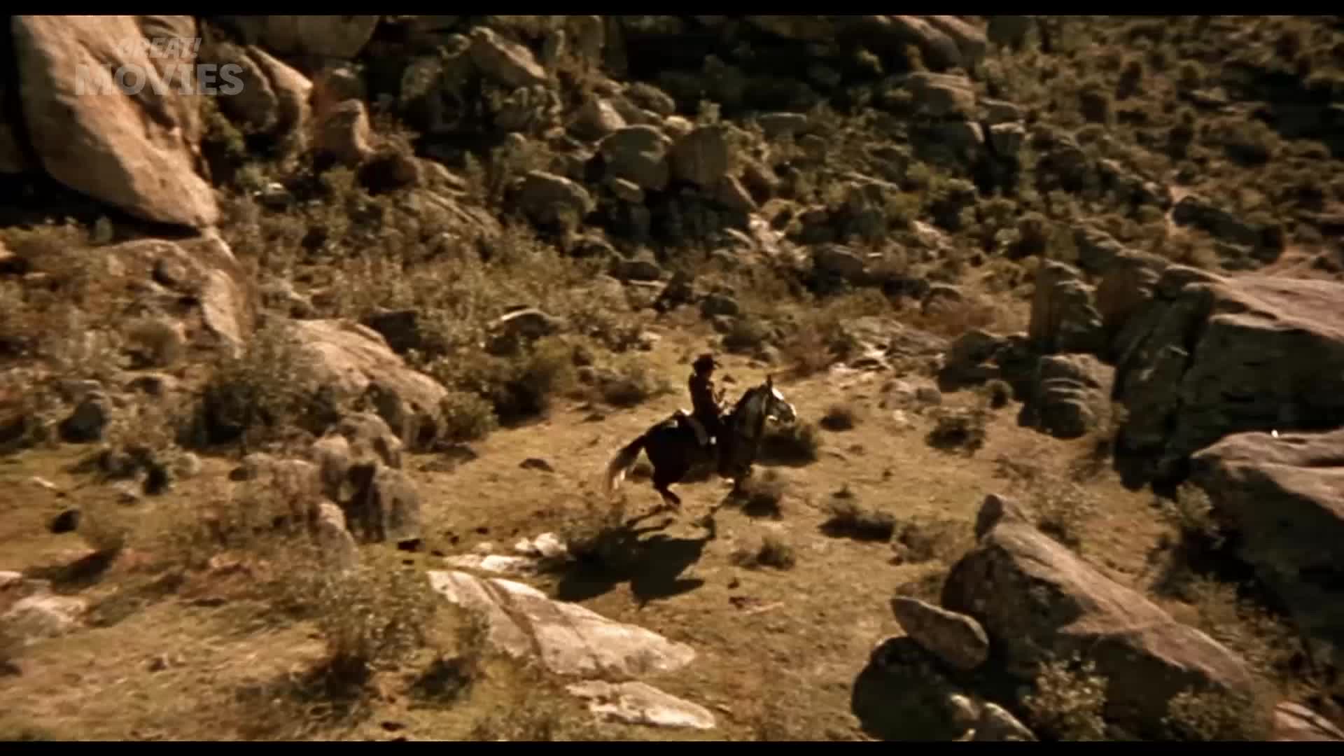 A lone rider on a dark horse gallops across a rocky, scrub-covered hillside. The horse's hooves kick up dust as it navigates the uneven terrain.