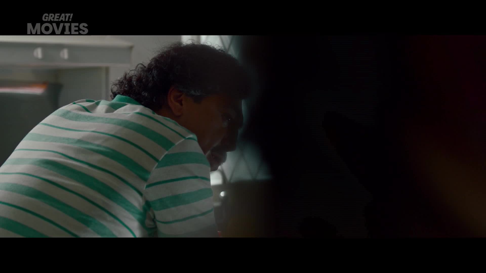A man in a striped polo shirt leans forward, his head bowed. The dim lighting and the GREAT! MOVIES logo suggest this is a scene from a film.