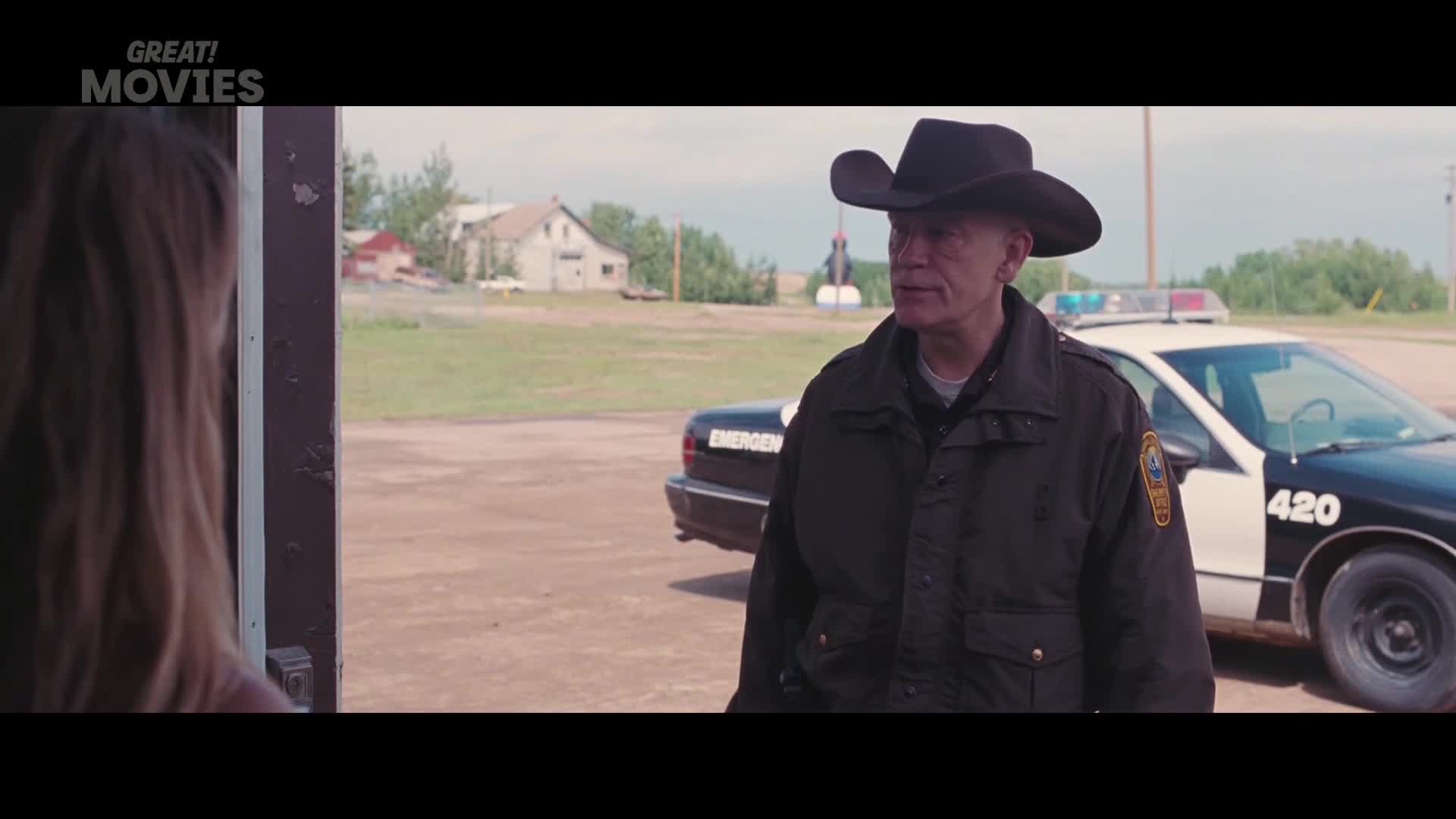 A man in a cowboy hat and a dark jacket stands near a police car. He's looking towards someone off-camera, his expression serious.