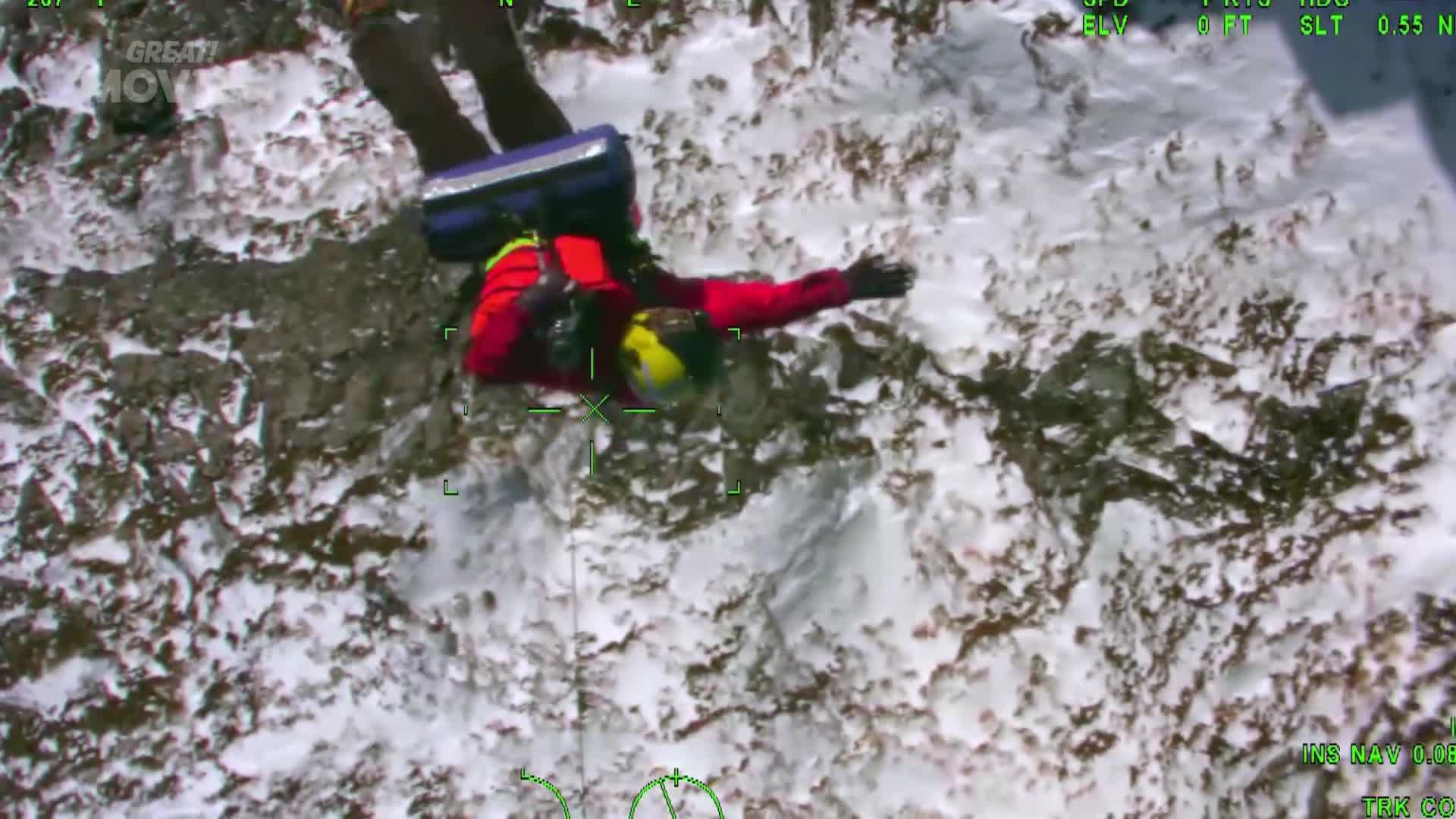 A rescuer in a red jacket and yellow helmet is being lowered down a steep, snow-covered mountainside. They are dangling from a cable, their arms outstretched as they descend towards the rocky terrain below.