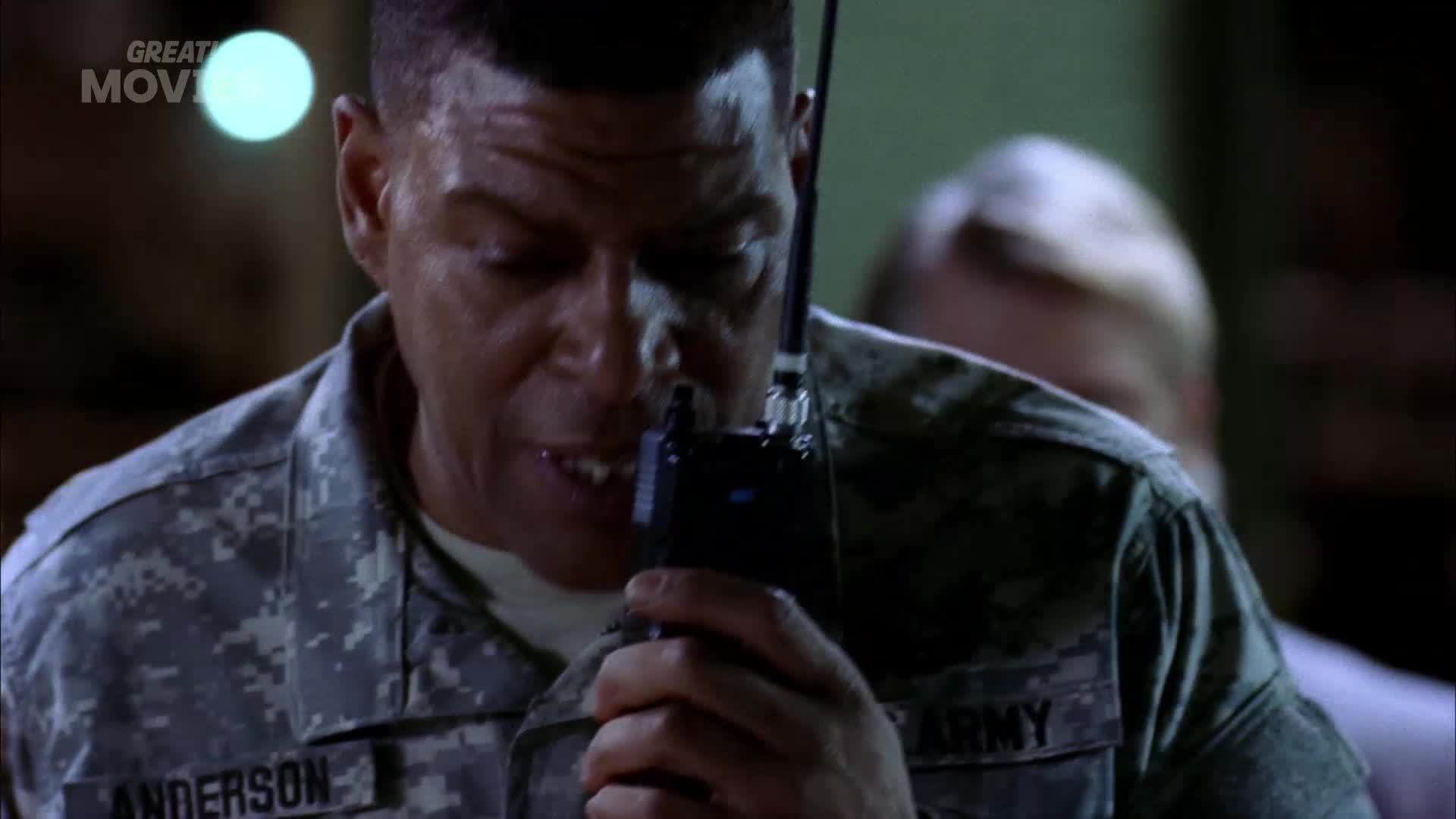 A soldier in camouflage speaks urgently into a radio. Another soldier, blurred and fair-haired, stands behind him, their shoulder visible.