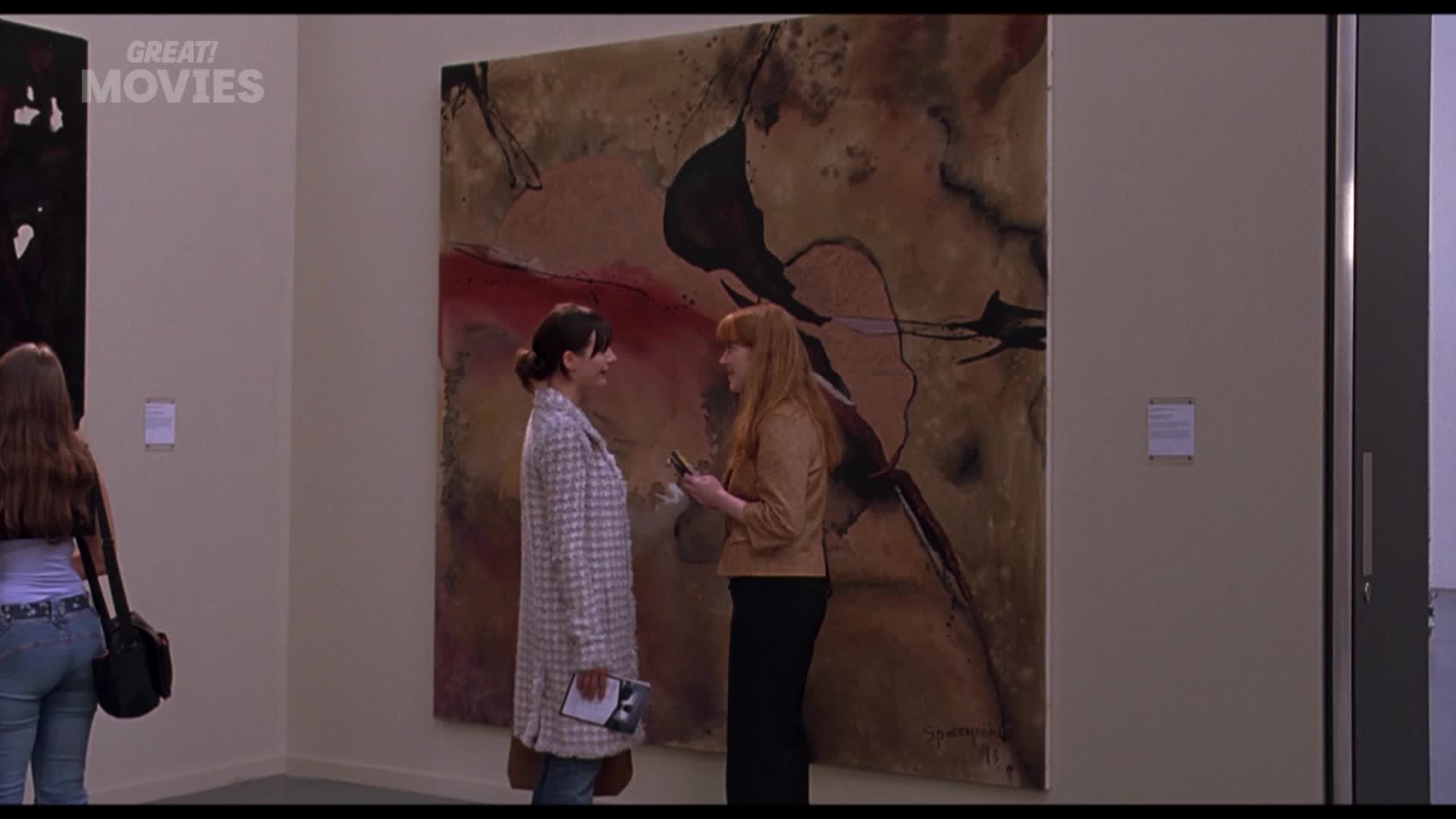 Two women stand before a large abstract painting, one holding a phone. Another woman, seen from behind, walks past a black canvas on the left.