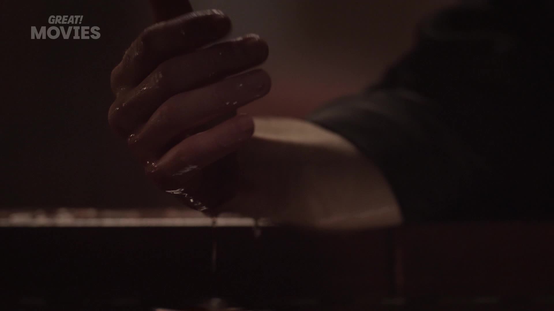A hand, slick with liquid, drips onto a dark surface. A faint "GREAT! MOVIES" logo hovers in the corner.