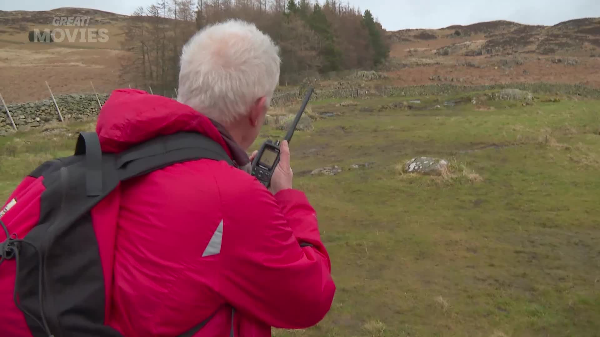 A man in a bright red jacket holds a radio to his ear, scanning the rugged, grassy hills of the UK. His gaze is fixed on something in the distance, as if waiting for a signal.