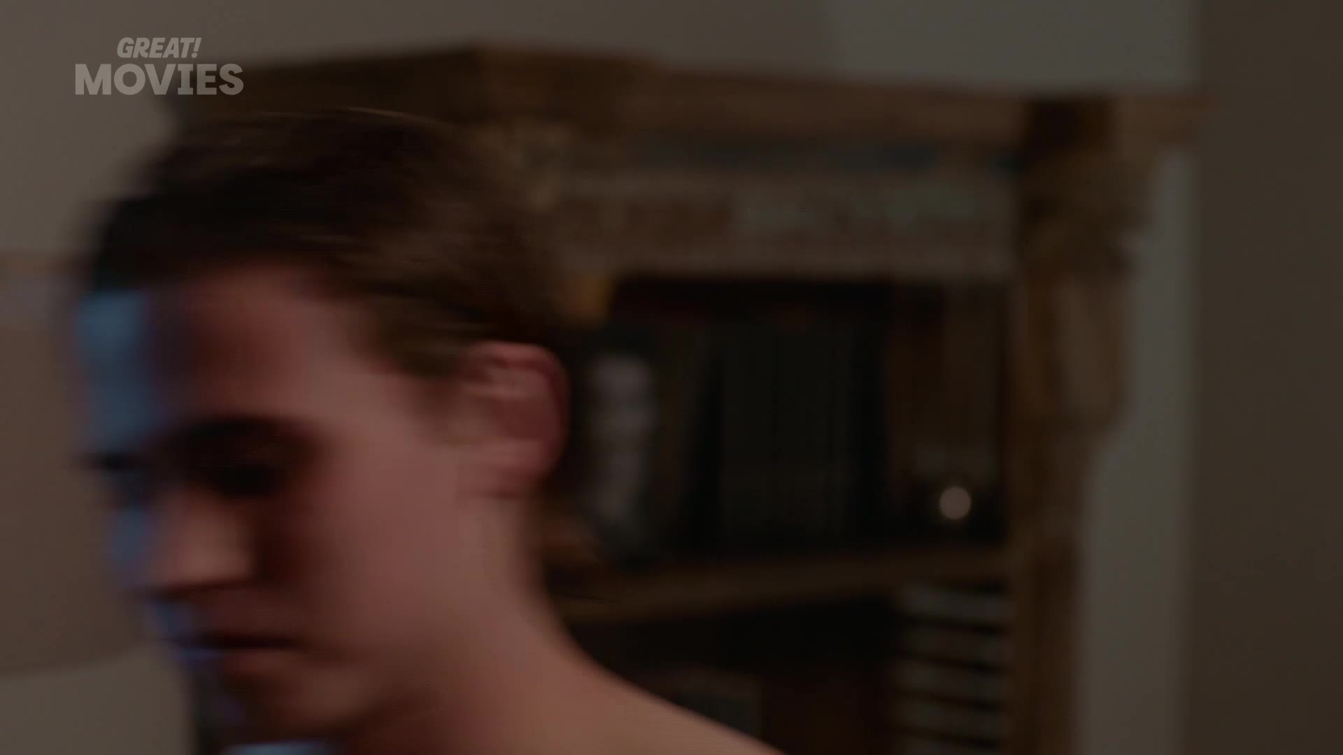 A young man's head turns sharply, his dark hair a blur against the dim background. He's in a room with what looks like a fireplace and bookshelves, a scene you might catch on GREAT! Movies.