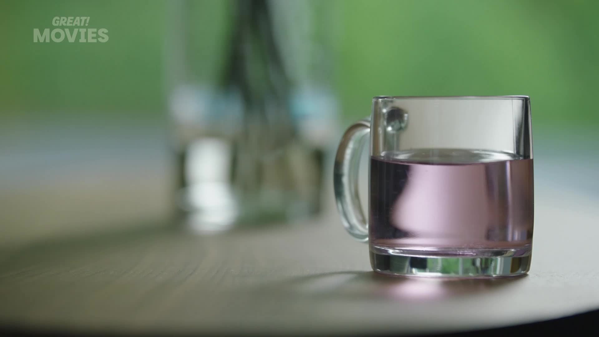 A clear glass mug, half-filled with a pale purple liquid, sits on a wooden surface. Behind it, a blurred background of green foliage suggests an outdoor setting, perhaps a garden on a pleasant afternoon.