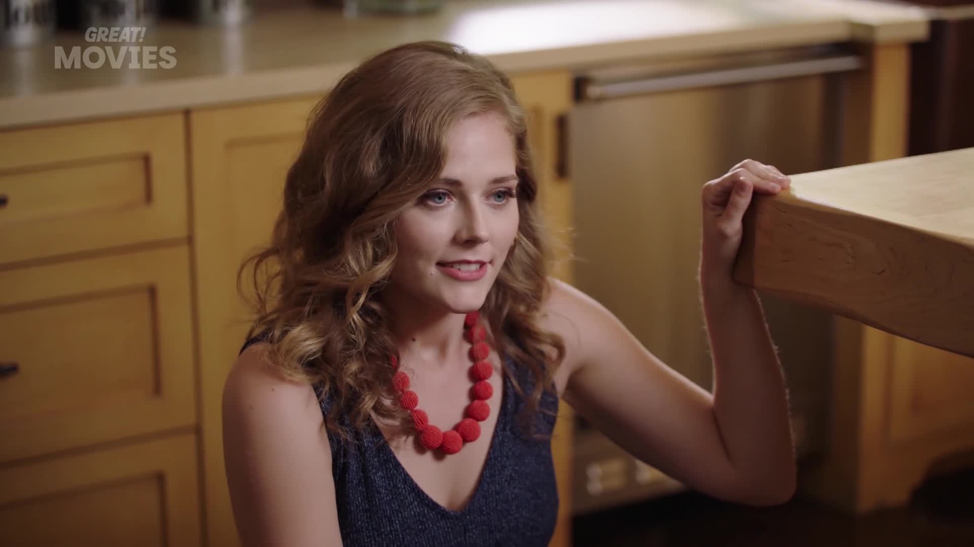 A young woman with curly blonde hair, wearing a dark blue dress and a striking red beaded necklace, leans forward. Her hand rests on the edge of a light-coloured wooden surface.