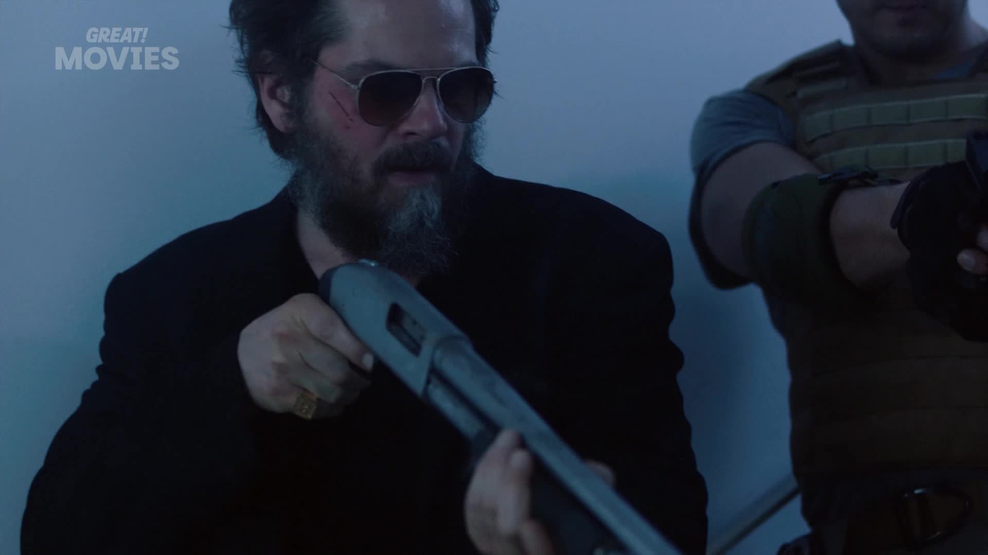 A man with sunglasses and a beard grips a shotgun, his face tense. Another man in tactical gear stands beside him, holding a firearm.