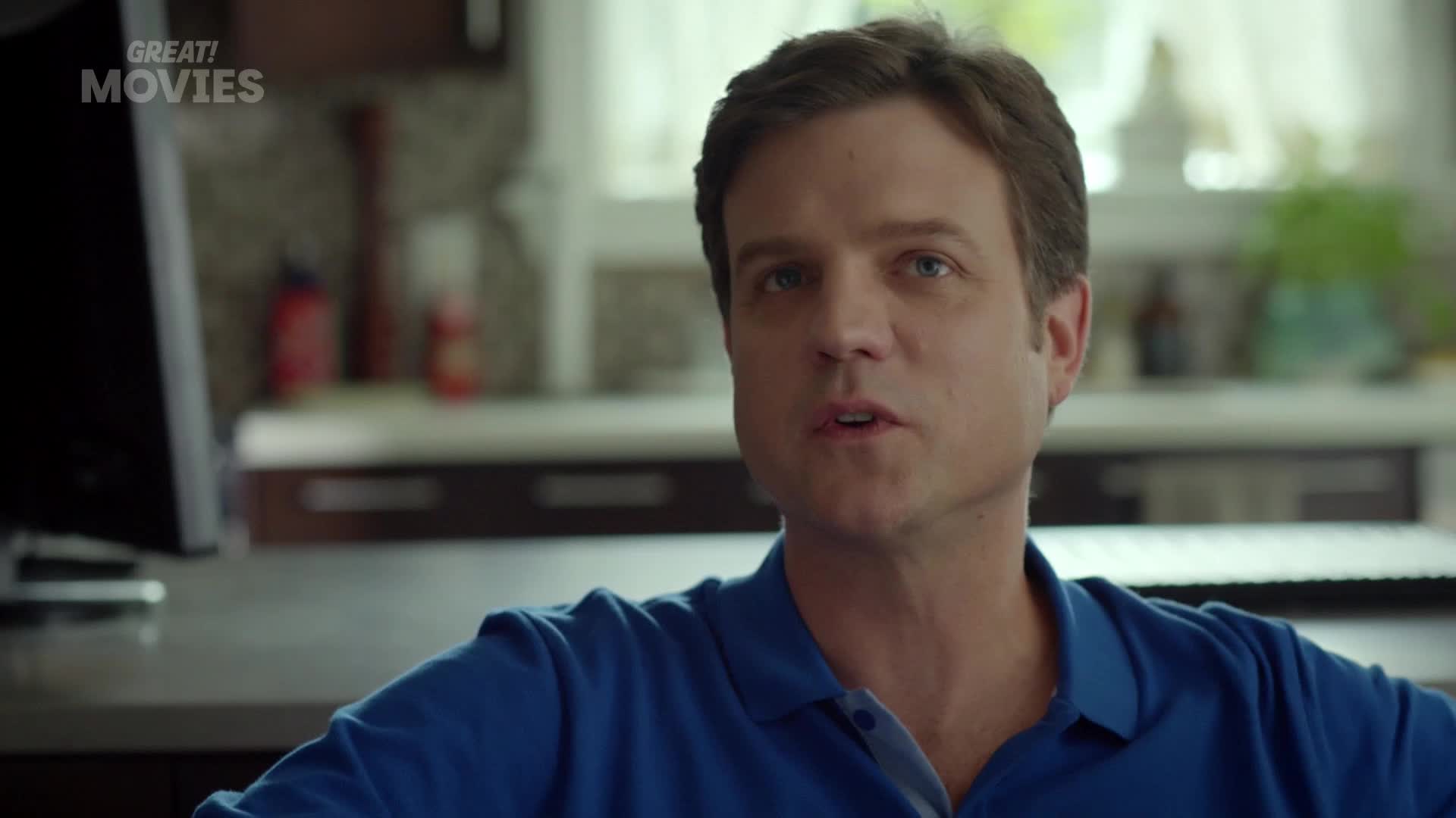 A man in a blue polo shirt speaks directly to me. Behind him, a kitchen counter holds a few bottles and a plant.