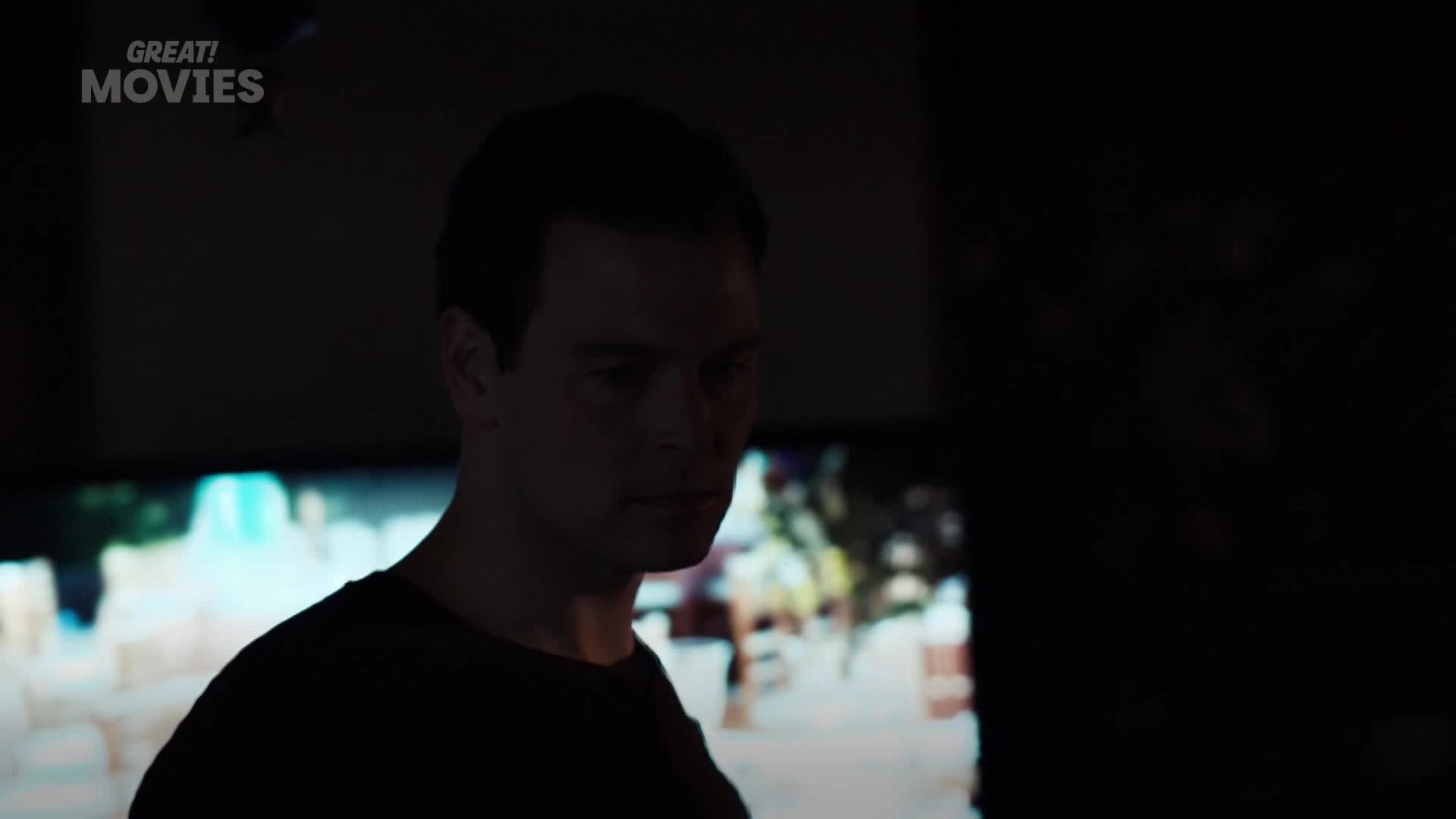 A man in a dark shirt stands in profile, his gaze fixed on something beyond the frame. Behind him, a television screen displays a bright, indistinct scene, perhaps a street in London.