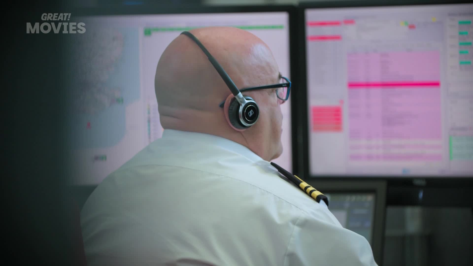 A man in a white shirt with gold epaulets is focused on multiple screens displaying data. He wears a headset, his gaze fixed on the glowing displays.