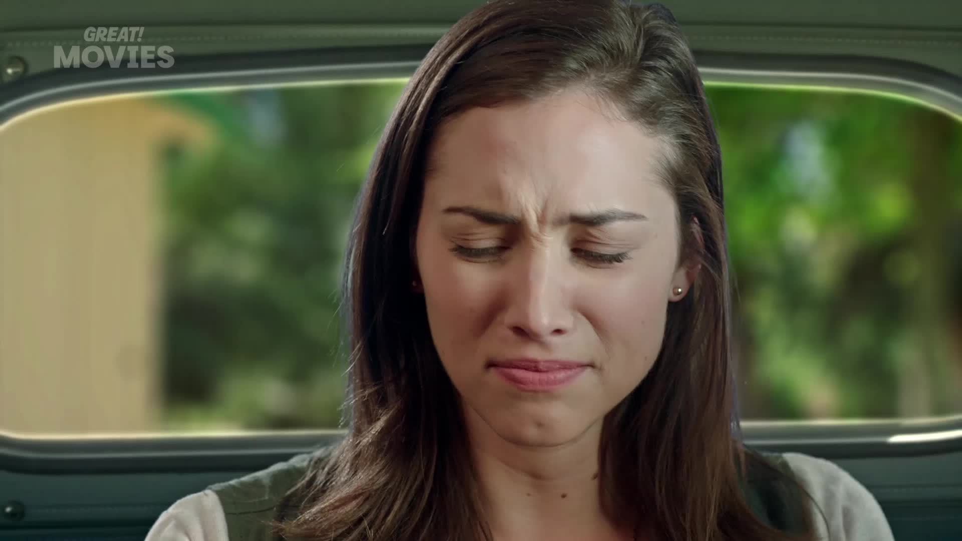 A young woman winces, her brow furrowed as she looks down. Outside the car window, trees blur past, suggesting a journey through the countryside.