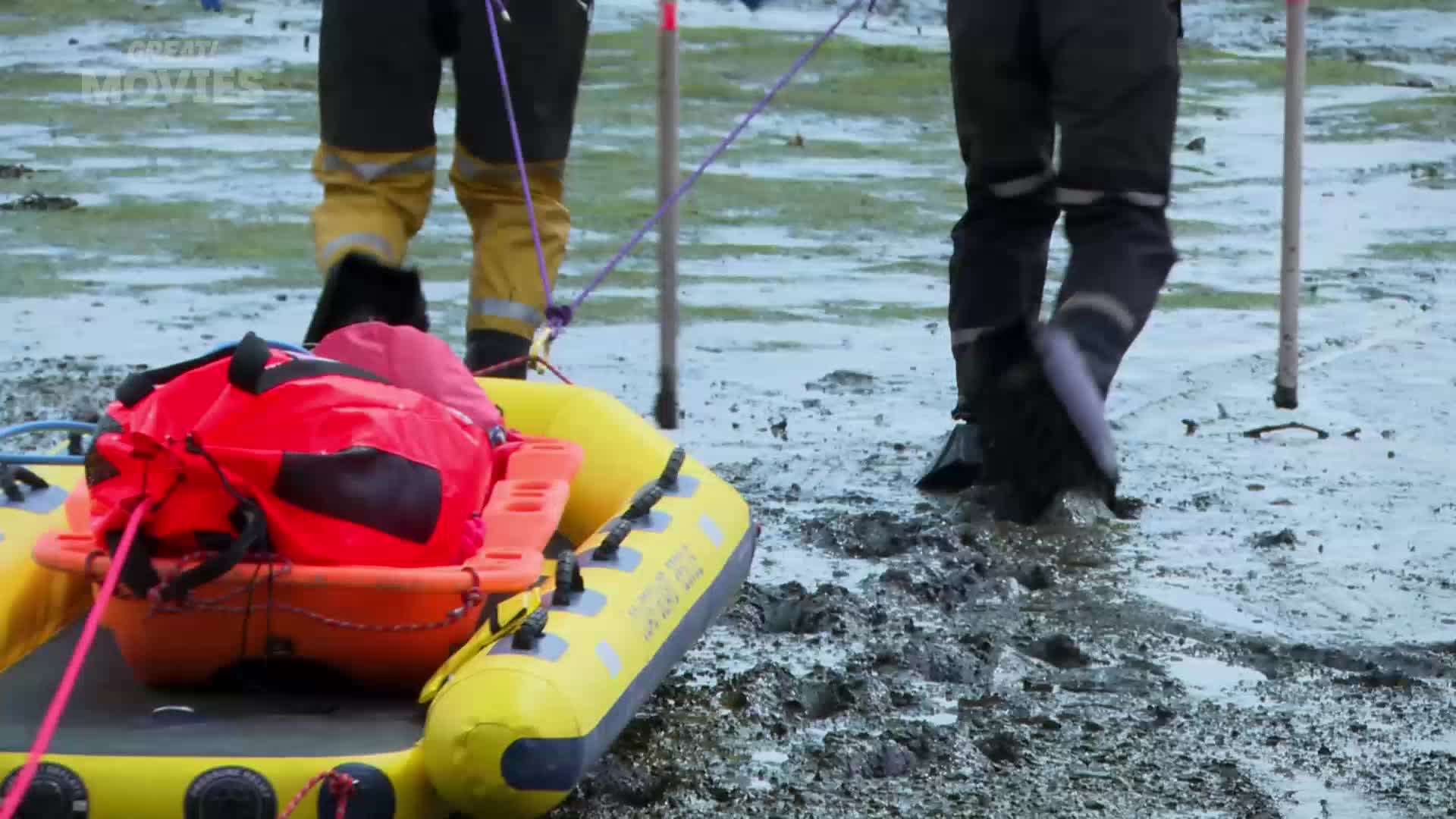 Two people in waterproof gear walk across a muddy, wet landscape, pulling a yellow inflatable raft laden with a bright red bag. The scene feels like it could be from a dramatic GREAT! movies production set in the UK.