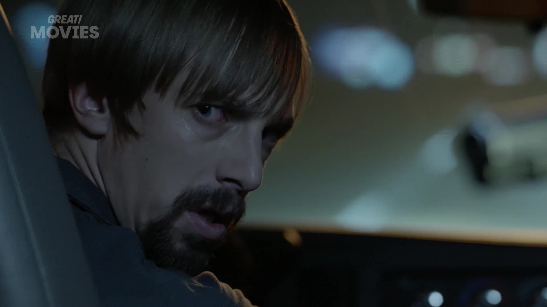 A man with a beard and a fringe glances over his shoulder, his eyes wide with alarm. The faint glow of streetlights passes by the car window.