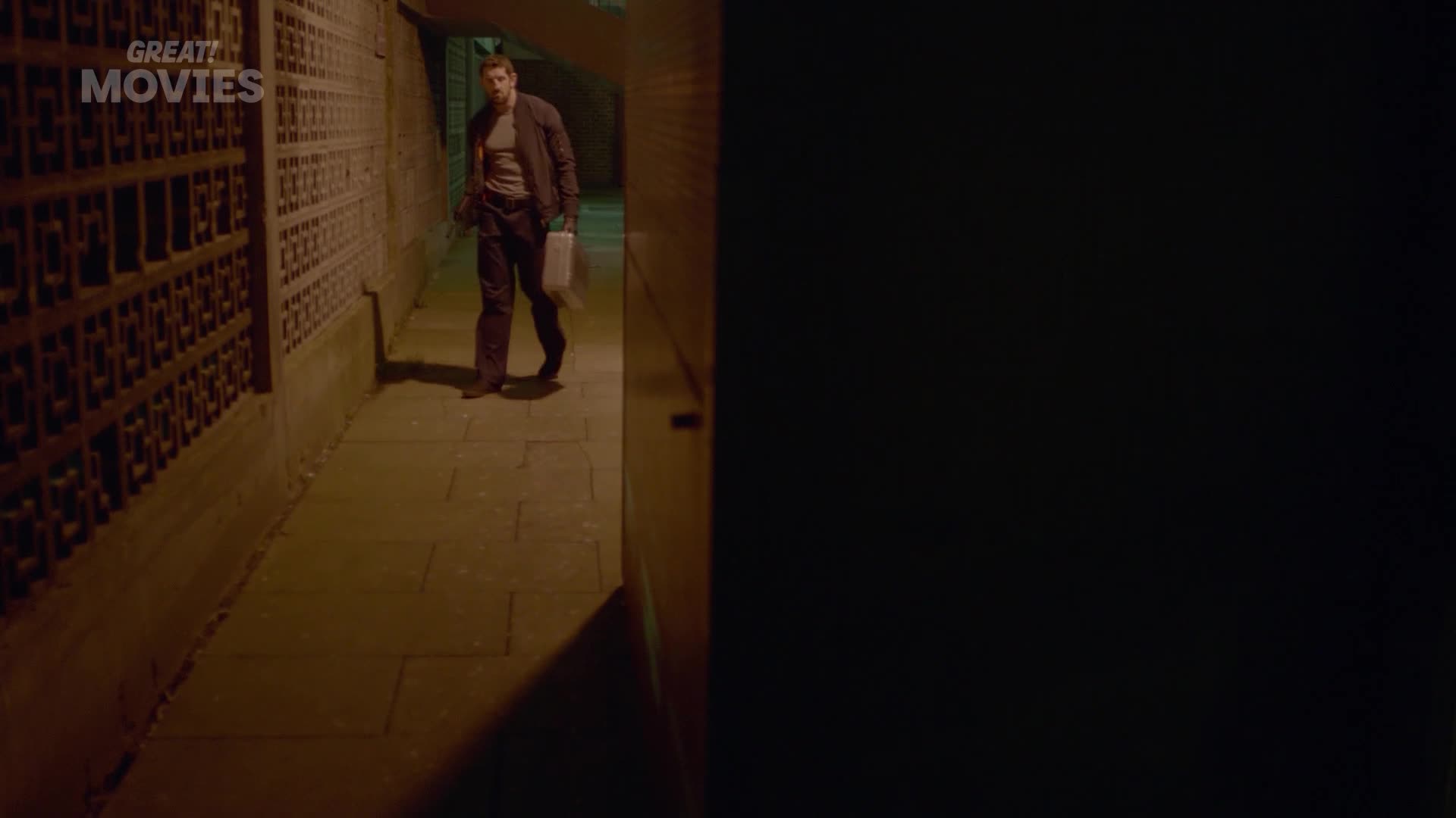 A man walks down a narrow alleyway at night, carrying a white bag. The brickwork on the left is illuminated by a dim light, casting long shadows.