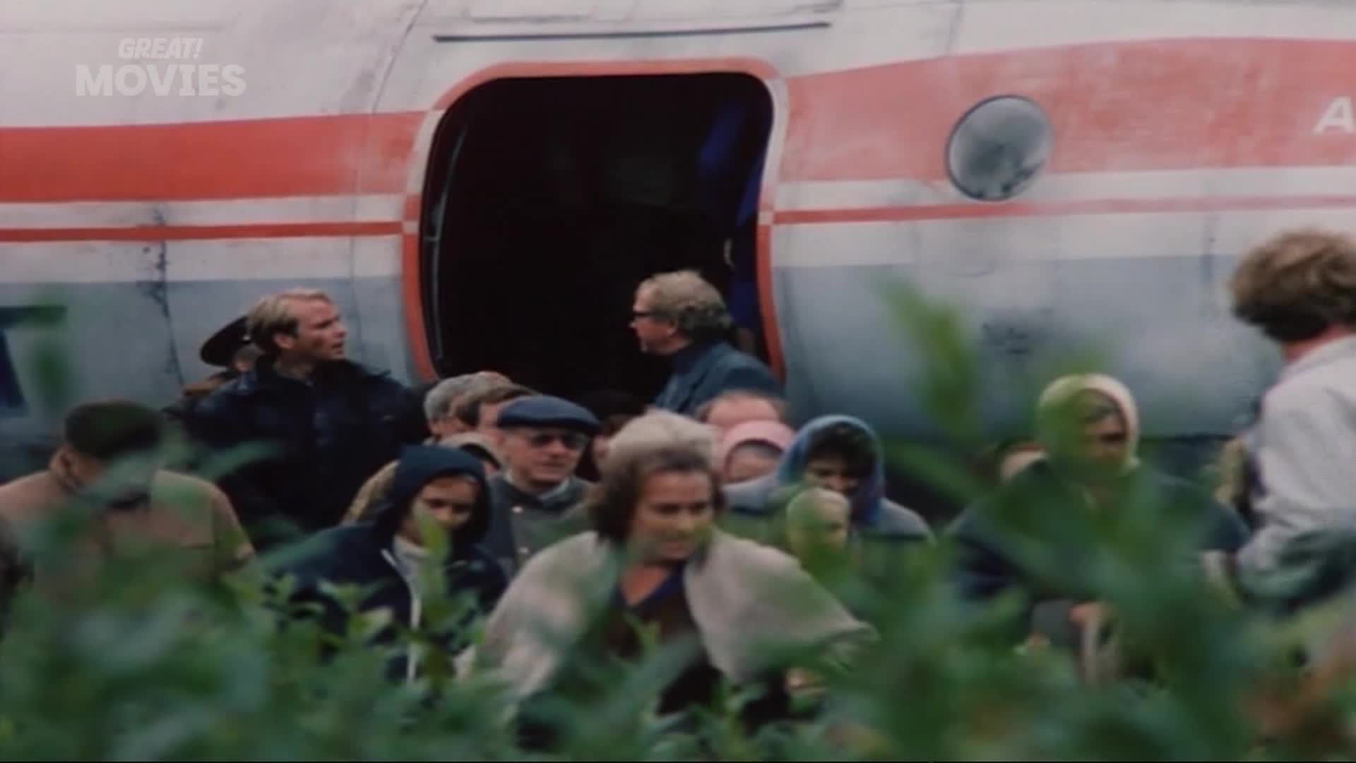 People are disembarking from a red and white aircraft, emerging from the open doorway into the overgrown landscape. A man in a dark jacket looks back towards the plane as a group of individuals, some with hoods pulled up, move forward through the greenery.