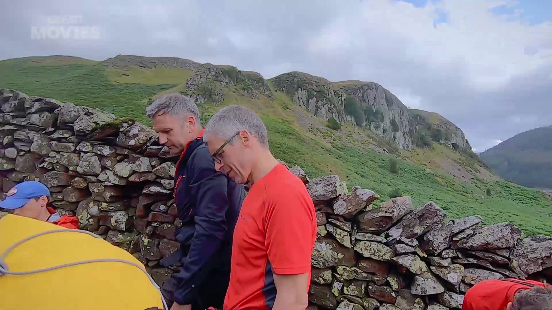 Two men in bright outdoor gear lean over a yellow object near a dry stone wall. The rugged, green fells of the Lake District rise behind them under a cloudy sky.