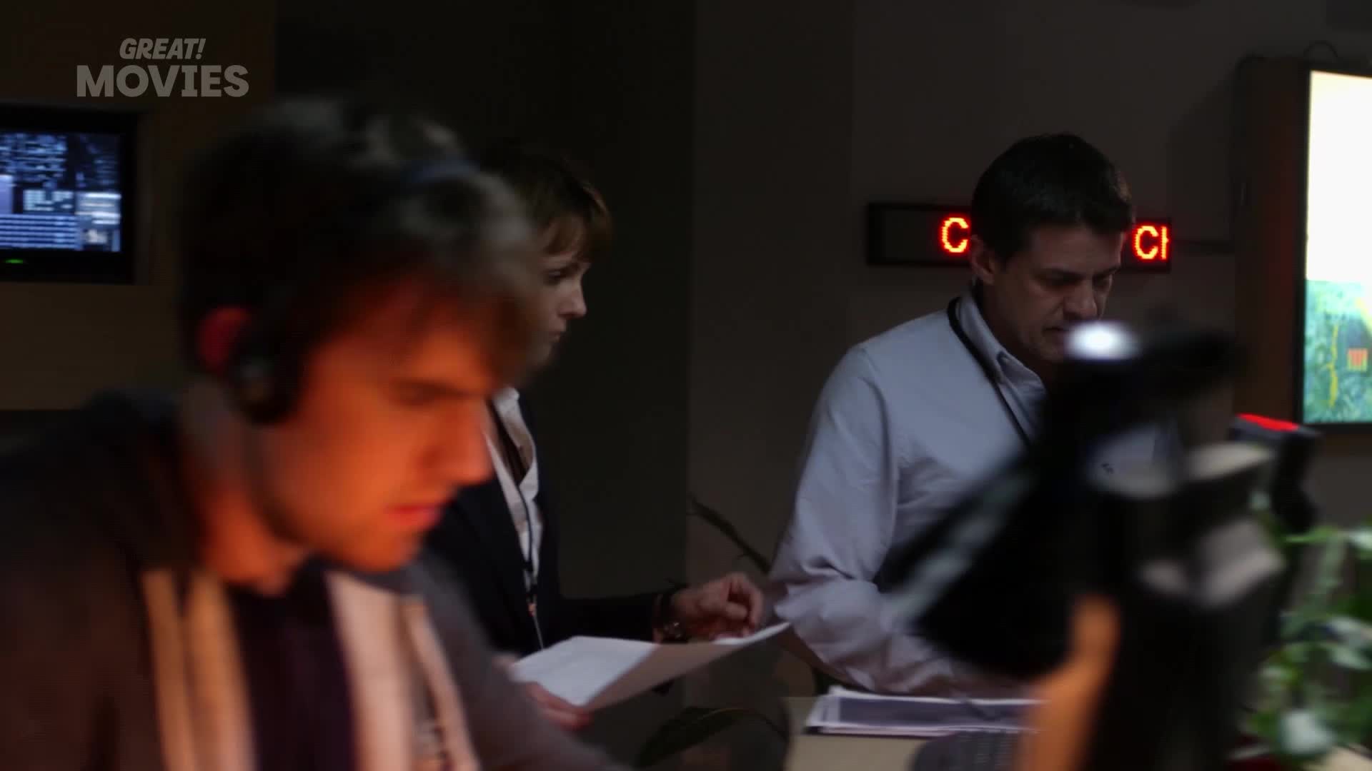 A young man with headphones leans over papers, while a woman beside him also focuses on documents. A man in a light blue shirt stands behind them, looking down at a desk.