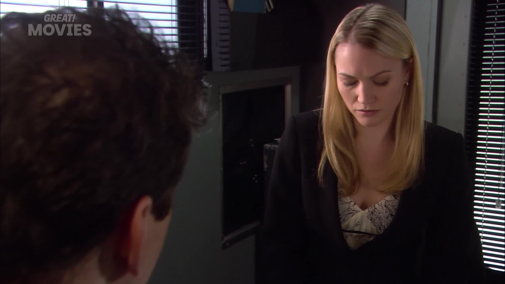 A woman in a black blazer looks down, her blonde hair falling forward. Beside her, a man with dark, curly hair is mostly out of frame.