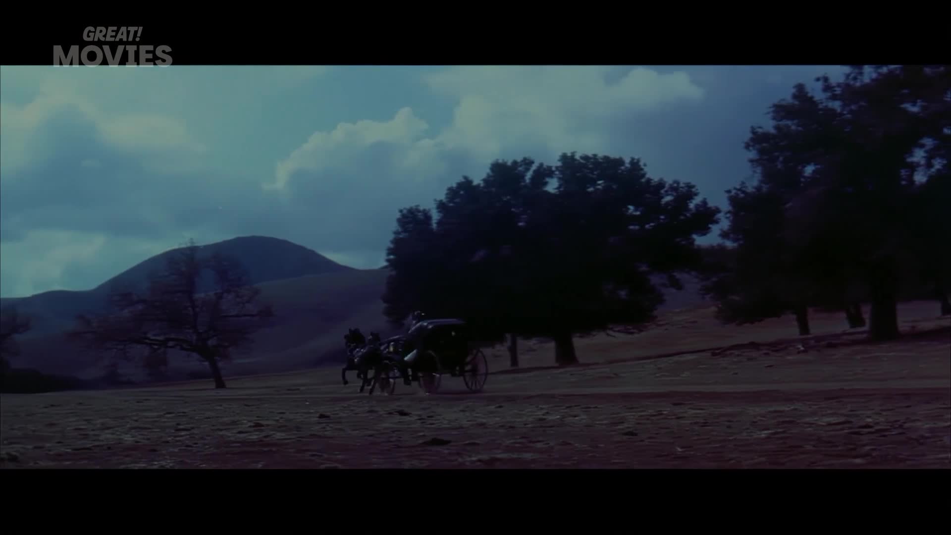 A horse-drawn carriage moves across a dusty field. The horses pull the dark vehicle with a steady gait, their hooves kicking up faint dust.