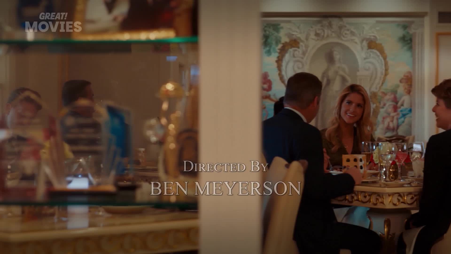 A woman with blonde hair smiles across a table at a man in a dark suit. The opulent room, reminiscent of a scene from a GREAT! Movies production, features ornate wallpaper and a crystal chandelier.