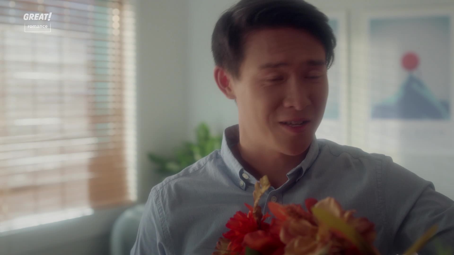 A man in a blue shirt holds a bouquet of red and orange flowers, his eyes closed as if he's trying not to cry. The scene feels intimate, like a moment from a GREAT! romance film.
