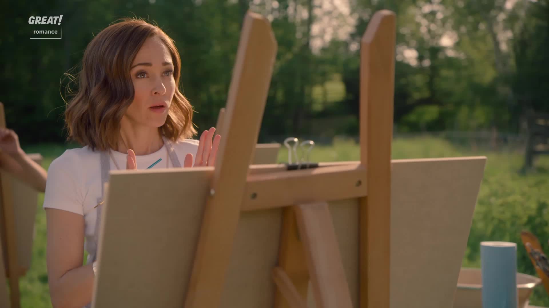 A woman in a white t-shirt and apron gestures with her hands, looking surprised. Behind her, a wooden easel holds a blank canvas, and the scene is set outdoors.
