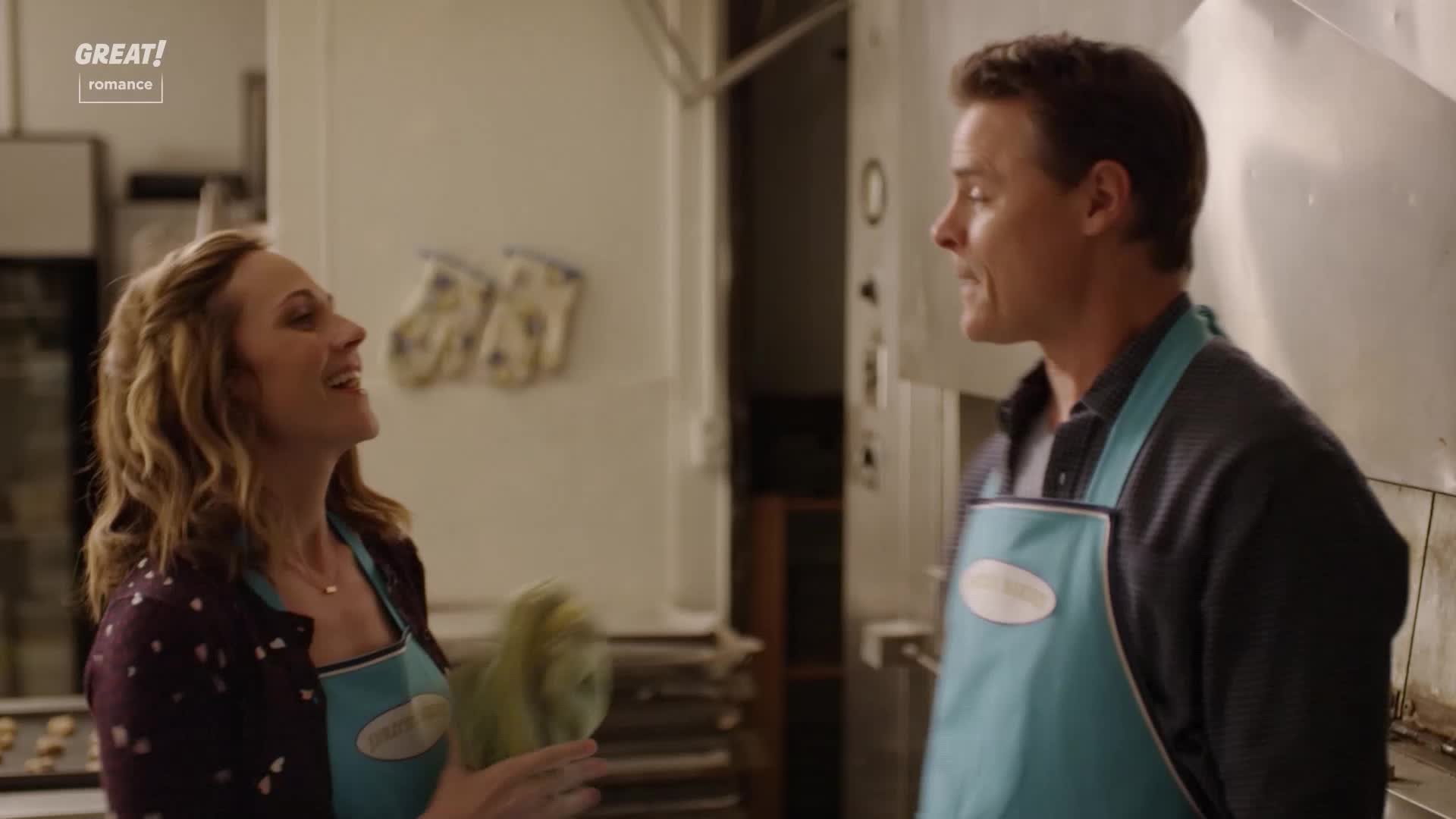 The woman is laughing, holding something green and fluffy in her hand. The man in the apron is talking, his expression engaged.
