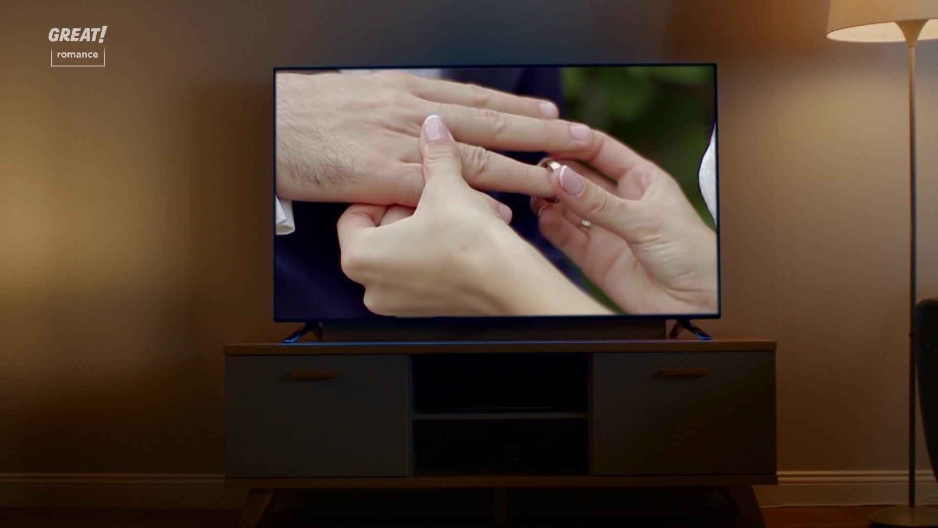 A hand gently slides a gold ring onto another hand's finger. The scene, playing on the television, looks like a moment from a GREAT! romance, perhaps set in the United Kingdom.
