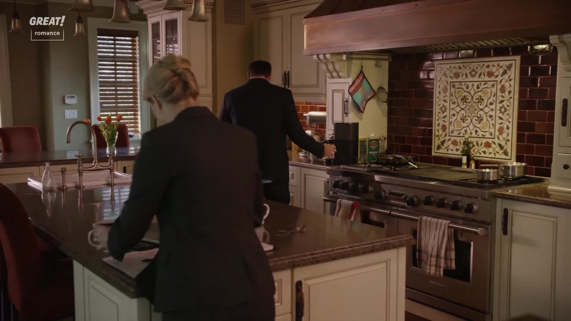 A woman in a dark suit sits at a kitchen island, looking at papers. Across the room, a man in a similar suit reaches for a coffee machine.
